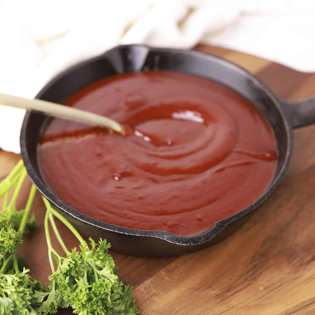 A skillet filled with Homemade BBQ Sauce, displayed on a wooden board with greens and a spoon in it.