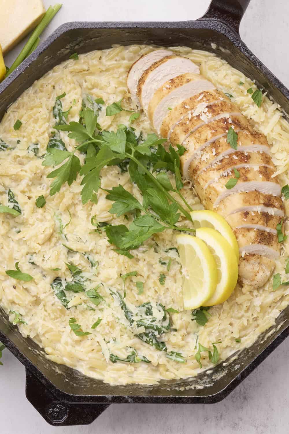 An overhead view of the Lemon Chicken Orzo Pasta in a skillet topped with cooked, sliced chicken breast, greens and lemon wedges.