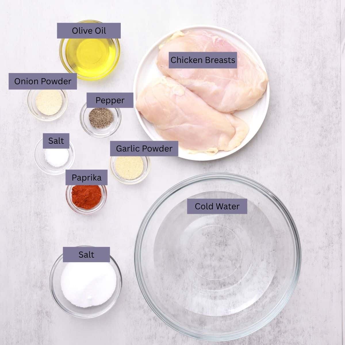 A visual display of the ingredients needed for the Skillet Chicken Breast recipe - chicken breasts, olive oil, onion powder, pepper, salt, garlic powder, paprika, water and more salt for brining - each in their own bowl.