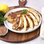 A whole and sliced Cast Iron Skillet Chicken Breast displayed on a white plate sitting on a wooden board and displayed with greens and a lemon.