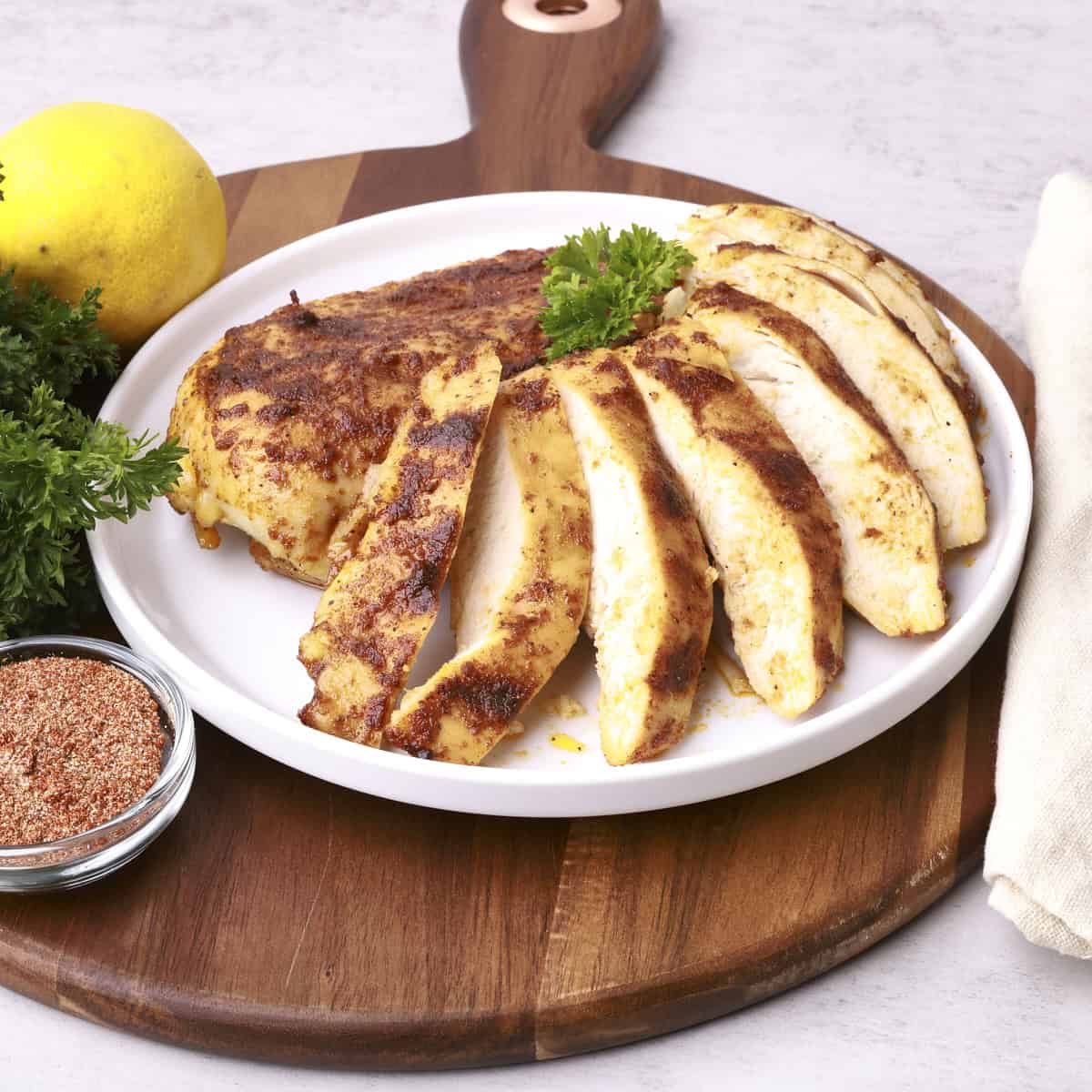 A whole and sliced Cast Iron Skillet Chicken Breast displayed on a white plate sitting on a wooden board and displayed with greens and a lemon.